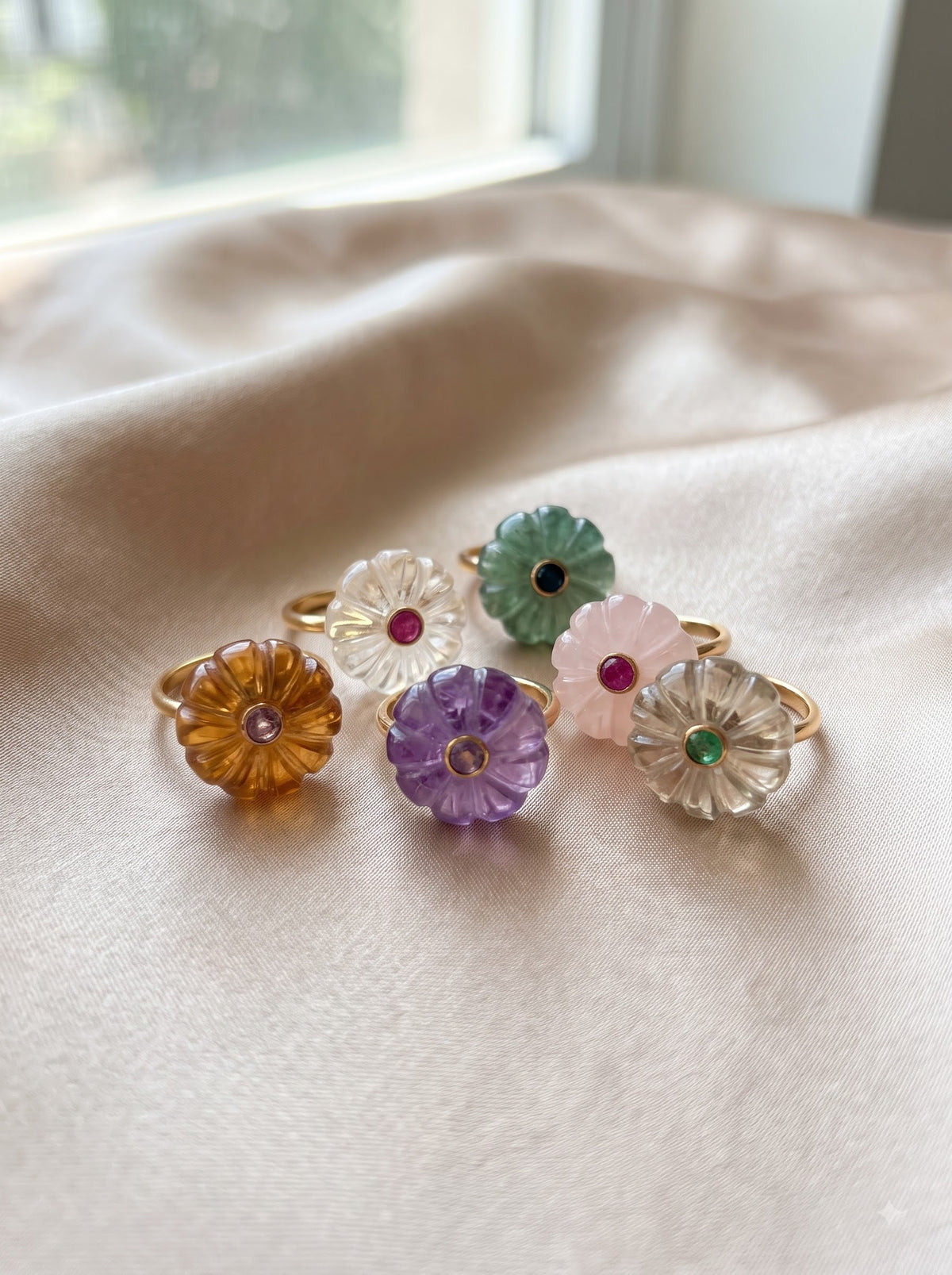 Five flower-shaped rings with gold-tone bands and translucent petals in amber, purple, white, mint green, and blush pink, each set with a small colored stone, arranged on a soft beige fabric surface near a window.