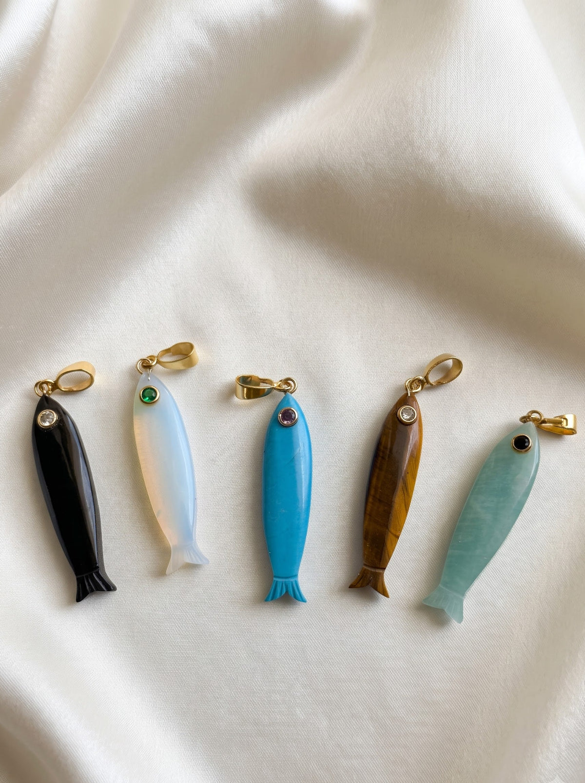 Five small fish-shaped jewelry charms or pendants arranged in a row on draped white fabric. The fish are made from different materials and colors: black, milky white, bright blue, brown wood grain, and light aqua