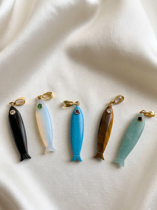 Five small fish-shaped jewelry charms or pendants arranged in a row on draped white fabric. The fish are made from different materials and colors: black, milky white, bright blue, brown wood grain, and light aqua