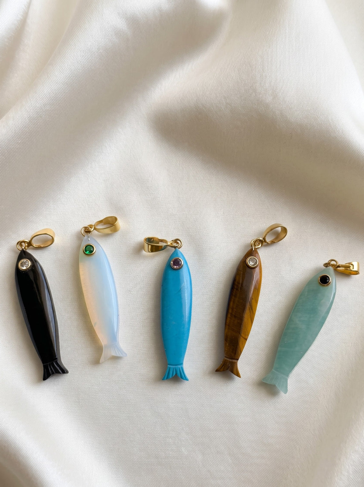Five small fish-shaped jewelry charms or pendants arranged in a row on draped white fabric. The fish are made from different materials and colors: black, milky white, bright blue, brown wood grain, and light aqua