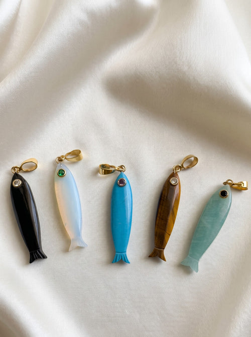 Five small fish-shaped jewelry charms or pendants arranged in a row on draped white fabric. The fish are made from different materials and colors: black, milky white, bright blue, brown wood grain, and light aqua